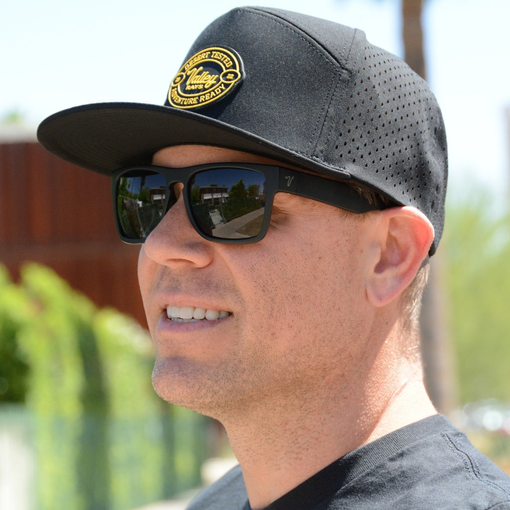 A cheerful man sporting a black trucker cap with a 'Valley Rays Desert Tested' patch and reflective polarized sunglasses, with urban scenery reflected in the lenses