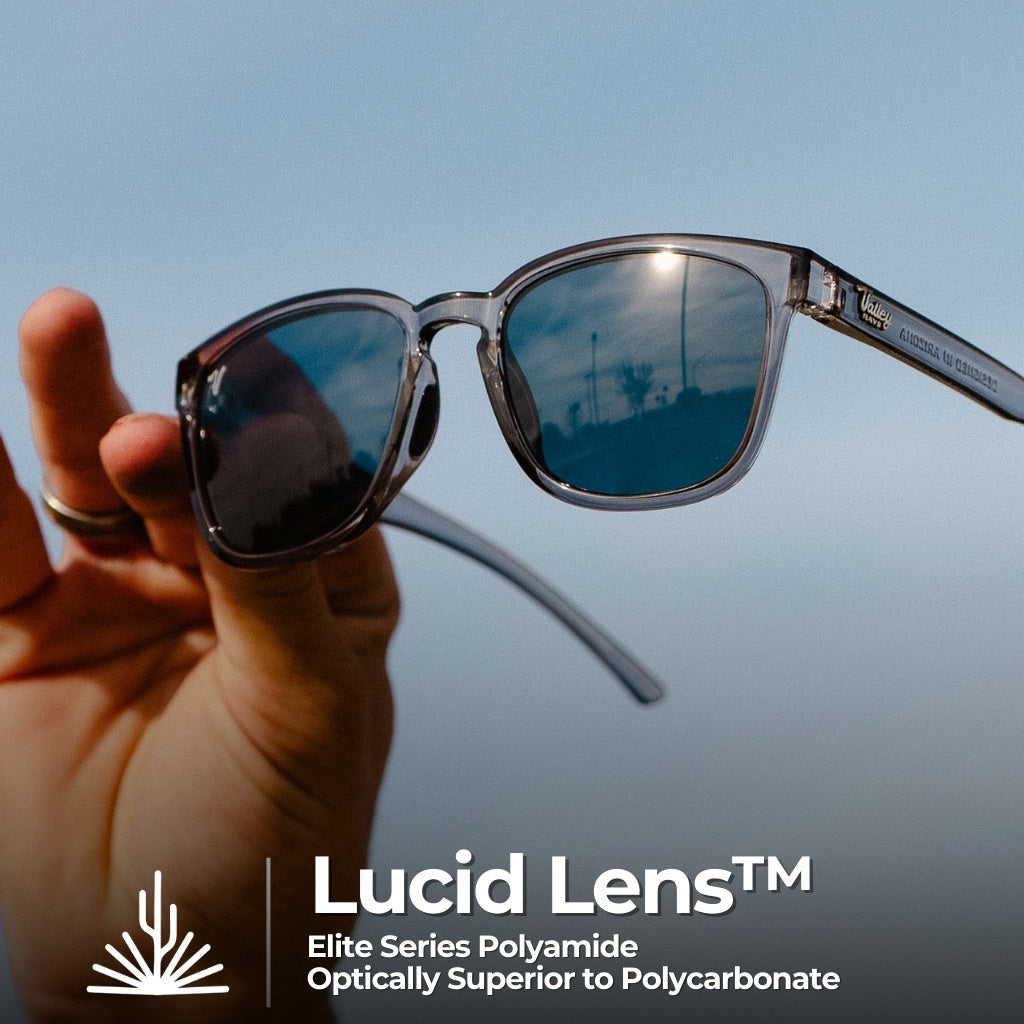 A man's hand elegantly holds a pair of stylish polarized sunglasses with reflective lenses, capturing the blue sky and clouds in their reflection, ideal for outdoor wear for men