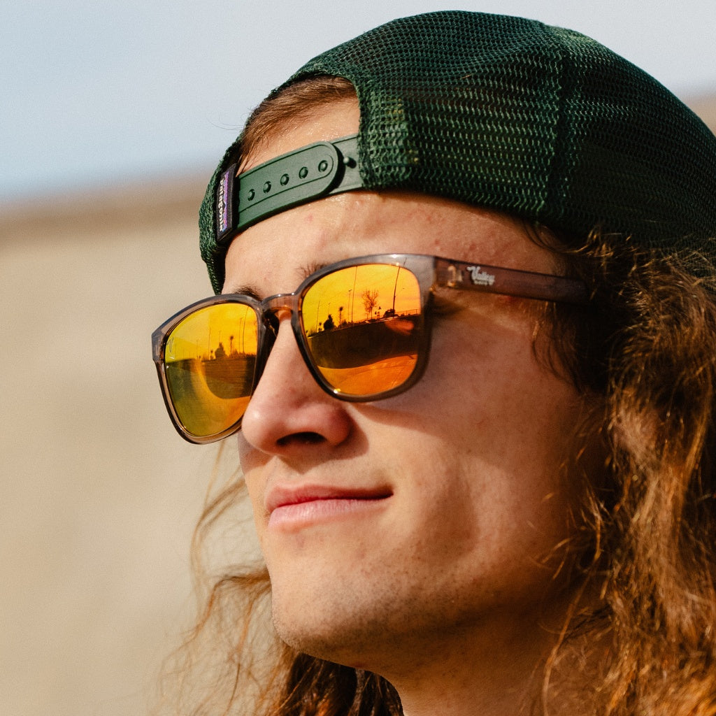 A smiling man with curly hair wearing a green backward baseball cap and yellow-tinted polarized sunglasses that reflect a sunset, exuding a relaxed, fashionable vibe